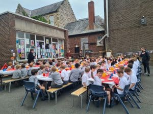 Outdoor school event with children seated at tables in a courtyard setting.