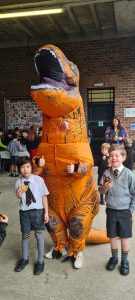 Colorful dinosaur mascot at Inchmarlo event, with children enjoying outdoor activities.
