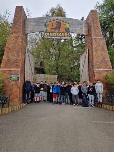 Group of visitors at Dinosaur theme park entrance with dinosaur sign and lush greenery, ideal for family outings.