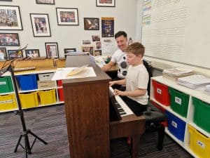 A teacher helps a student play piano in a classroom setting.