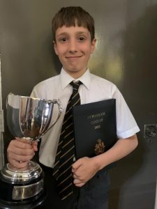 Young boy in school uniform holding a trophy and a book, celebrating academic success.