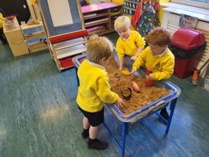 Three children playing in Inchmarlo nursery sensory table with sand and toys.