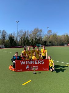 Group of young field hockey players celebrating with a winners' banner on a sports field.
