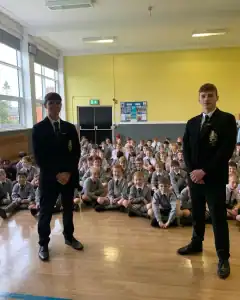 Students seated in assembly hall, two boys standing in front, engaging with students during school event.