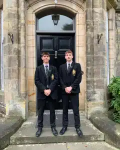 Two young men in formal suits standing at Inchmarlo estate entrance, historic stone building background.