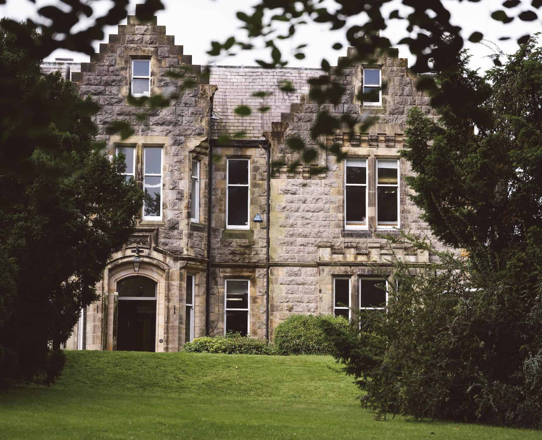 A picturesque stone-built Scottish manor surrounded by mature trees and well-maintained lawns.
