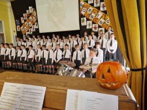 School children singing on stage at Inchmarlo, festive event with Halloween decorations and pumpkin.