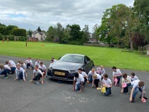 Students sitting on ground outside Inchmarlo School, green landscape in background, educational outdoor activity.