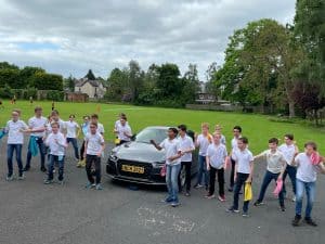 Group of young people standing outdoors with a car on a grassy area, enjoying a daytime event.