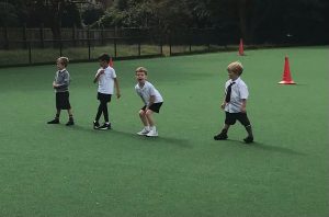 Children playing on a green school playground with cones in the background.