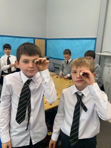 Two young boys in school uniform holding magnifying glasses in classroom.