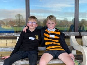 Two young boys sitting on a bench, smiling outdoors at Inchmarlo School with a clear sky background.