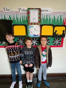 Three children in festive sweaters standing in front of a holiday-themed display at Inchmarlo.