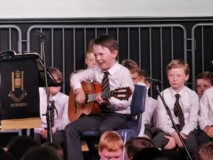 Young boy playing guitar at Inchmarlo school performance, surrounded by classmates.