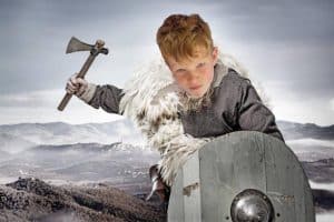 A young boy dressed as a Viking with an axe and shield on a rugged landscape.