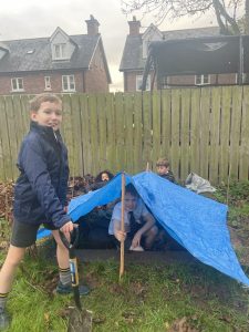 Children building a small shelter with a blue tarp in a garden.