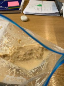 Close-up of a baking mixture in a clear plastic bag on a wooden table, with papers and a blue pen in the background.