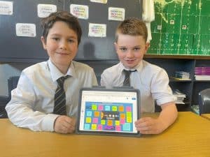 Two boys in school uniforms holding a tablet with colorful app, in a classroom.